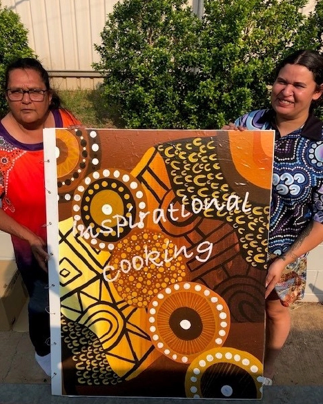 Two people holding a large Aboriginal artwork with the words Inspirational Cooking